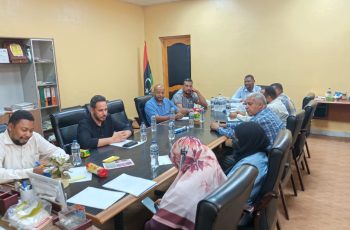 A business meeting around a long conference table with ten people in an office, several water bottles, papers, and a Libyan flag in the background.