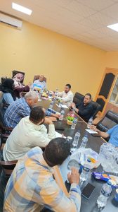Group of professionals seated around a long conference table in a yellow-walled meeting room, discussing and taking notes with water bottles and snacks on the table.