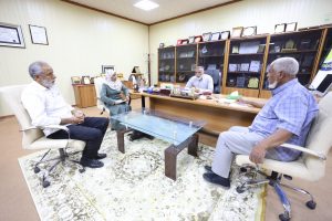 Four adults in a formal office seated around a glass coffee table for a meeting, with shelves of awards behind them.