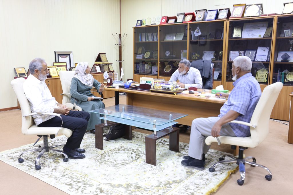 A group of four light-skinned adults seated around a glass meeting table in a wood-framed office, discussing papers with awards displayed behind them.