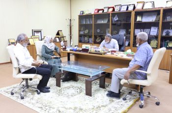 A group of four light-skinned adults seated around a glass meeting table in a wood-framed office, discussing papers with awards displayed behind them.