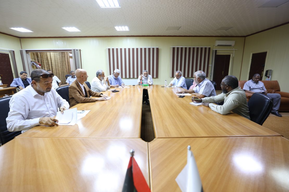 Group of men seated around a long wooden conference table in a meeting room, with small flags visible at the table foreground.