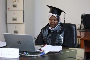 Woman in graduation gown and cap sits at a desk with an HP laptop, papers, and markers in an office setting