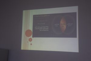 Lecture slide projected on a wall, showing a diagram with two overlapping circular shapes and an orange central lens, with Arabic labels and a small red page number '13' in the corner.