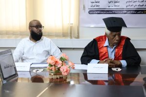 Two men sit at a conference table during a formal ceremony; one in a white shirt, the other in a black robe with red trim and cap, with flowers on the table behind a name card banner.