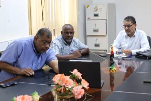 Three men in a meeting around a conference table, focused on a laptop; Libyan flag on the table and office in the background.