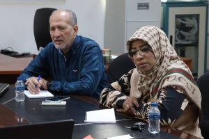 Two professionals sit at a conference table; man writes in a notebook while a woman in a patterned hijab listens and reviews documents, with a water bottle nearby.
