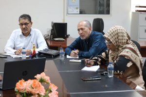 WhatsApp Image 2026-10-25 at 11.33.55 PM - جامعة سبها | Sebha University Three professionals sit around a conference table in an office: a man in a white shirt on the left, a man in a blue shirt in the center, and a woman wearing a patterned hijab on the right, all taking notes with laptops, notebooks, and water bottles nearby.