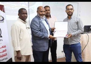 - جامعة سبها | Sebha University Award presentation: one man hands a certificate to another as three onlookers stand nearby in a formal setting.
