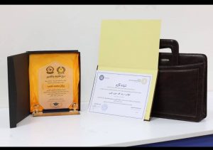 Award plaque in an open black box with orange lining, placed next to a pale yellow certificate folder and a brown leather briefcase on a white table.