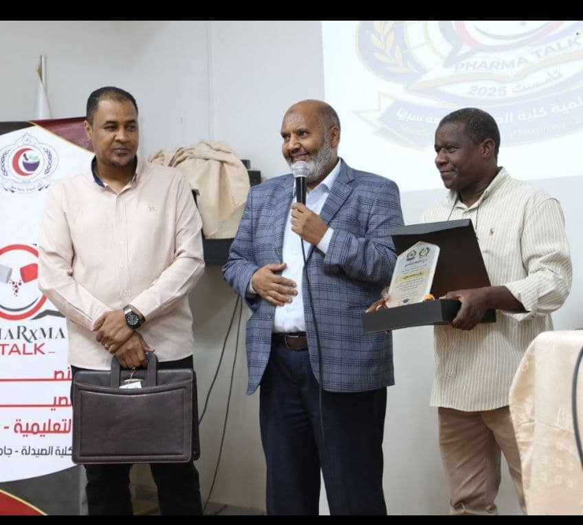 Three men at an awards ceremony; center man speaks into a microphone while the right man holds a plaque in a presentation box.