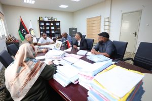 WhatsApp Image 2026-10-25 at 12.41.04 PM - جامعة سبها | Sebha University Group of people around a conference table discussing documents in a formal office setting with flags and awards in the background.