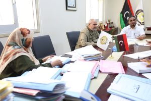 WhatsApp Image 2026-10-25 at 12.41.05 PM (1) - جامعة سبها | Sebha University Woman in a patterned hijab sits at a cluttered conference table with papers, facing two men in a meeting room; Libyan flags and trophies visible in the background.