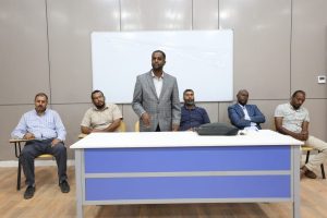 WhatsApp Image 2026-10-25 at 9.41.54 AM (1) - جامعة سبها | Sebha University Speaker in a gray plaid blazer addresses a panel of five men seated at a long table in a conference room, with a whiteboard behind them.