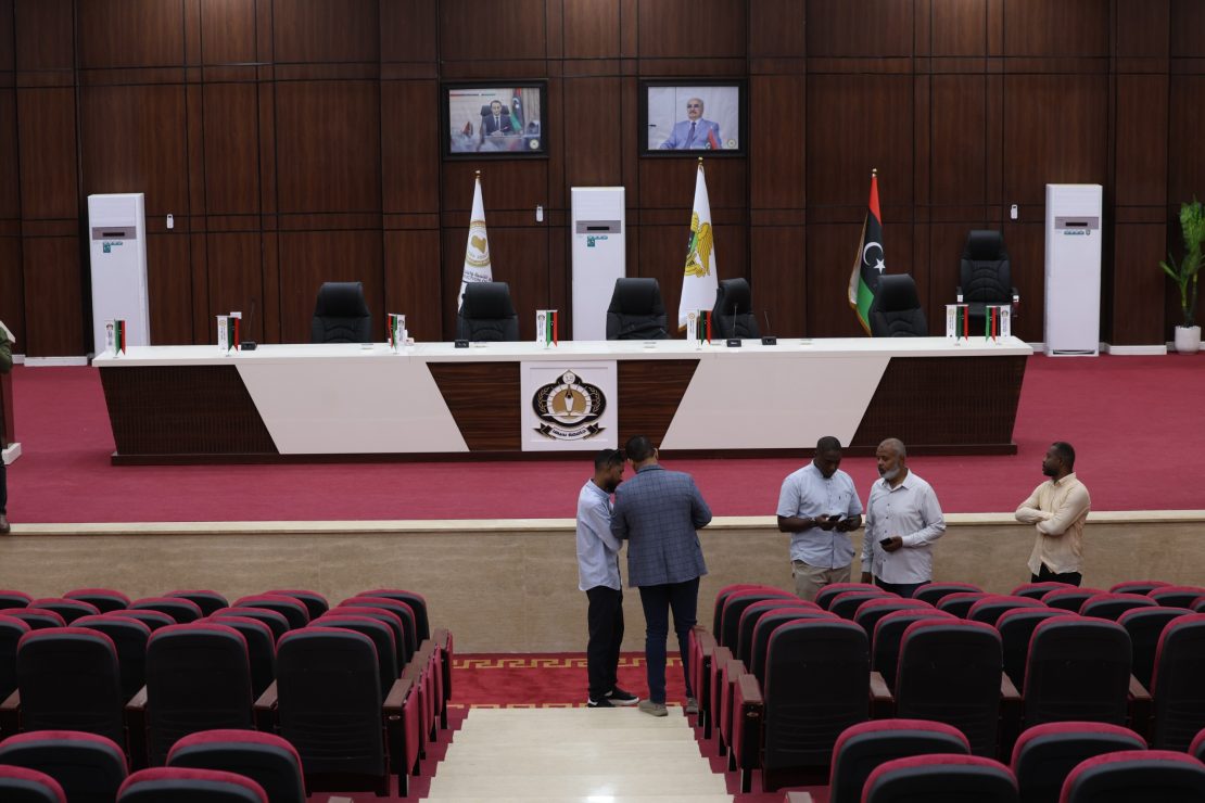 Conference hall with a long white desk on a red carpet, flags, and black executive chairs; a few men stand in front of the desks as audience seats fill the foreground.