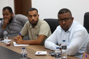 Three men sit around a conference table in a business meeting, with papers and pens in front of them and water bottles nearby.