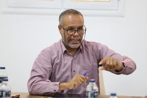 Older man with glasses and a striped pink shirt gestures while seated at a table during a meeting.