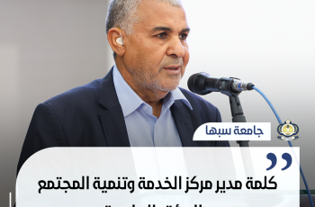 رجل يتحدث إلى الجمهور أمام ميكروفون على منصة جامعية، مع شعار جامعة سبها في الزاوية.